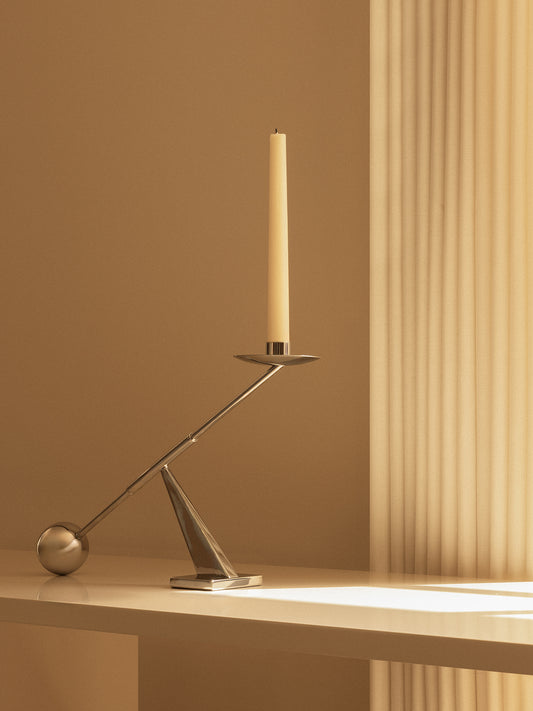 A geometrically designed interconnect candle holder in a polished steel finish, with a single candle placed on it, set against a warm-toned backdrop.